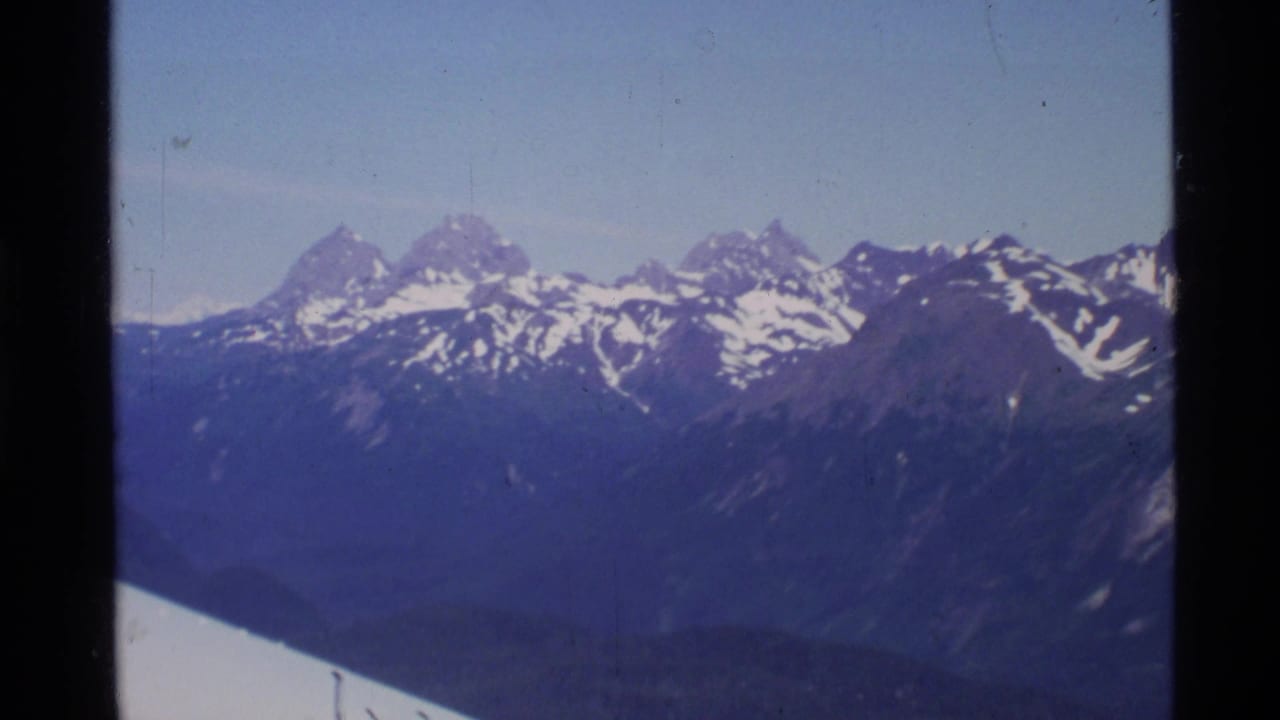 1977 Alaska 8mm aerial frame showing snowy mountain peaks and dark slopes through an aircraft window.
