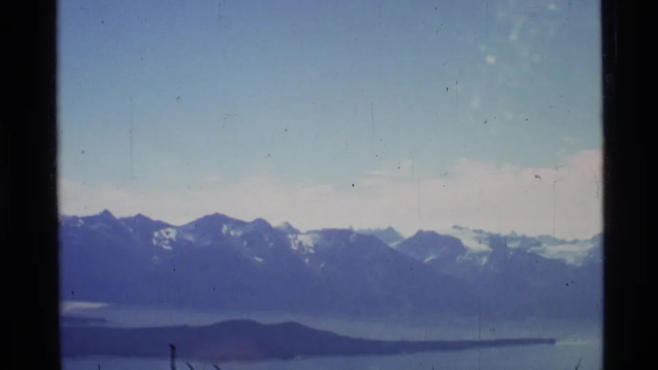 1977 Alaska 8mm aerial frame with a mountain skyline, broad water, and a dark low island under a pale sky.