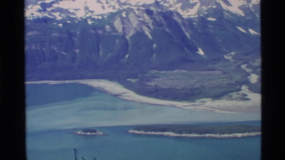 1977 Alaska aerial frame showing blue water, small forested islands, snowy mountains, and glacier-like ice below.