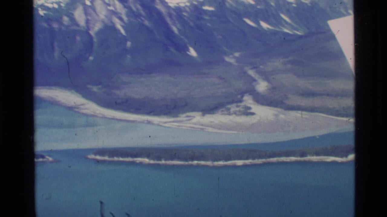 Aerial view of blue water, a narrow island, pale river flats, and dark mountains with snow patches.