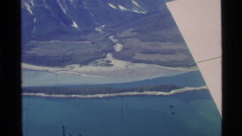 1977 Alaska home-movie frame showing turquoise coastal water, a long wooded island, pale river channels, and rugged mountain terrain.