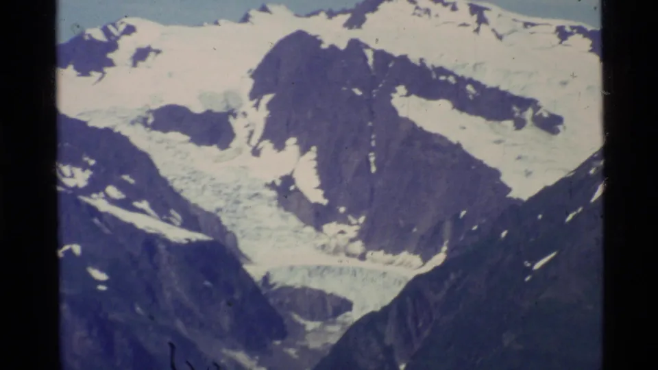 1977 Alaska 8mm aerial frame of rugged dark peaks with broad snowfields and glacier-like ice filling a mountain valley.