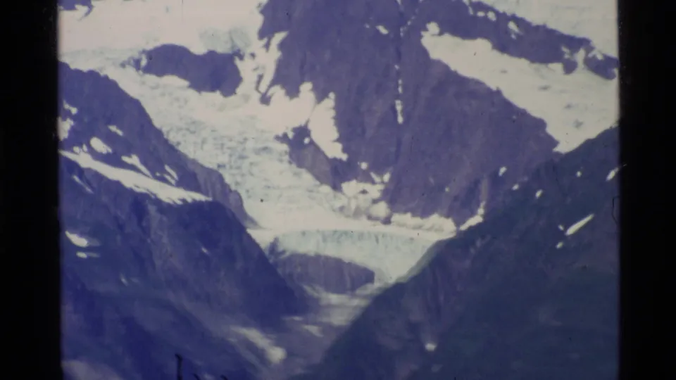 1977 Alaska aerial home-movie frame looking into a steep mountain valley with snow patches and glacier-like ice between dark ridges.