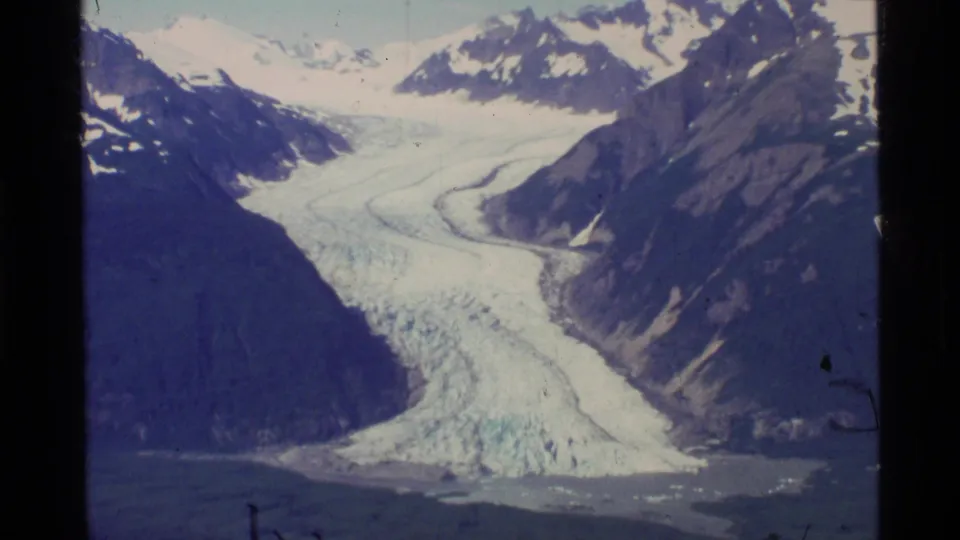 1977 Alaska 8mm frame of a wide glacier descending between rugged valley walls toward dark water below.