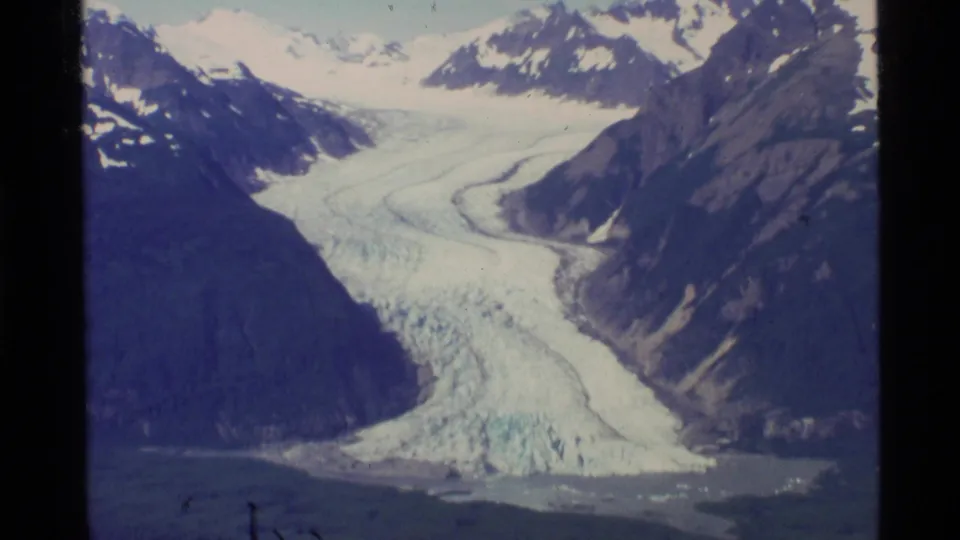 Vintage Alaska 8mm view of a glacier winding between steep mountain walls with snow fields beyond.