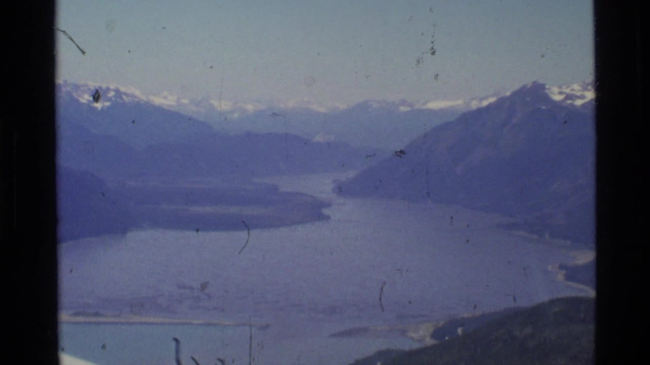 1977 Alaska aerial 8mm frame showing a broad lake or inlet framed by distant snow-capped mountains and dark film edges.