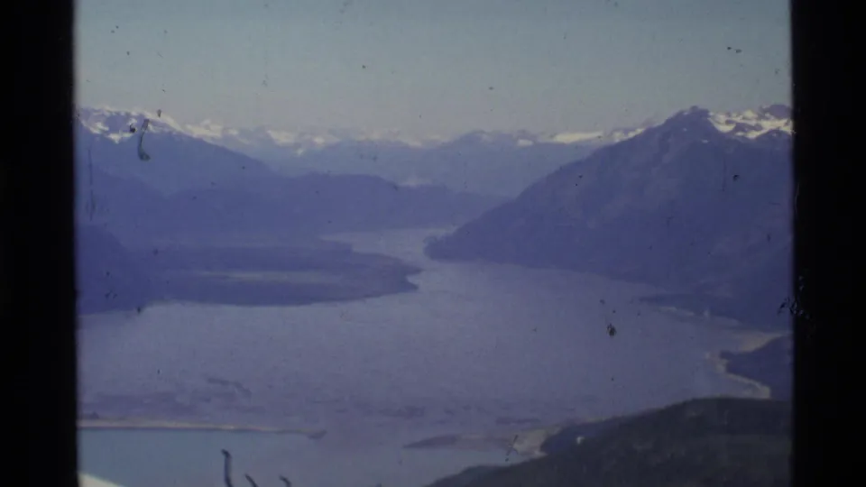 1977 Alaska aerial frame with blue lake water, valley terrain, and snow-capped mountain ridges in the distance.