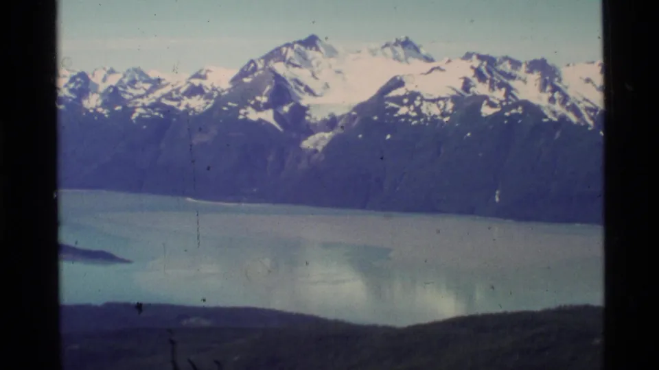 A 1977 Alaska 8mm aerial view of snow-capped peaks above a pale blue lake, with a dark ridge crossing the lower frame.
