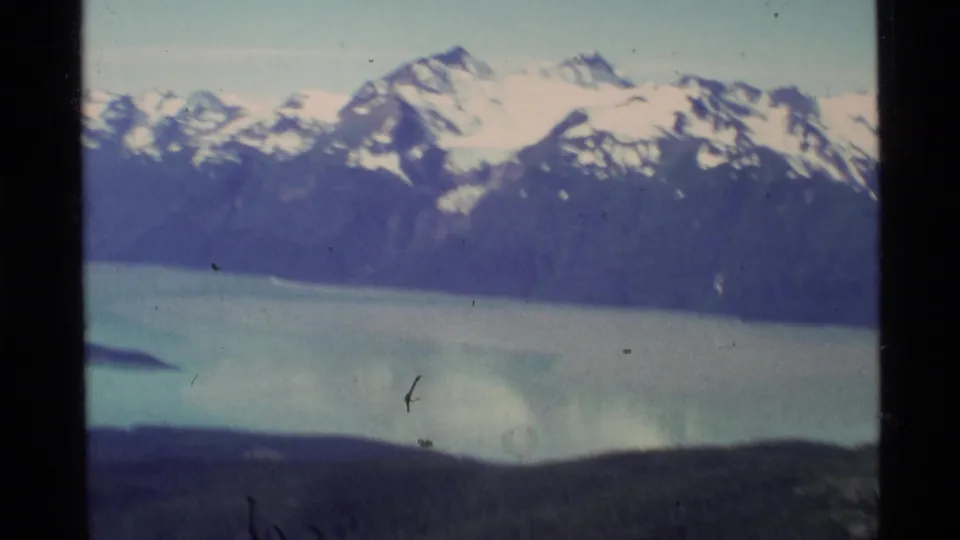 1977 Alaska aerial 8mm frame showing a mountain wall above milky blue lake water, with visible dust and soft focus.