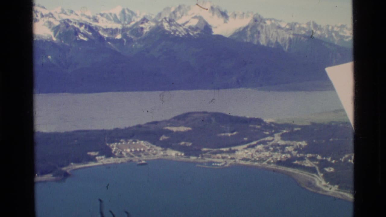 1977 8mm aerial view over coastal Alaska, showing shoreline buildings, blue water, forested hills, and distant snow-capped mountains.