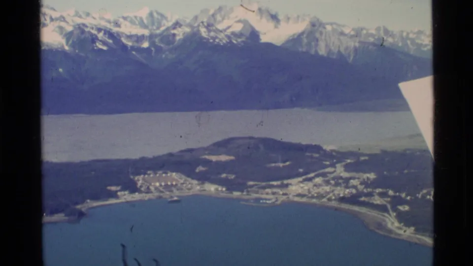1977 8mm aerial view over coastal Alaska, showing shoreline buildings, blue water, forested hills, and distant snow-capped mountains.