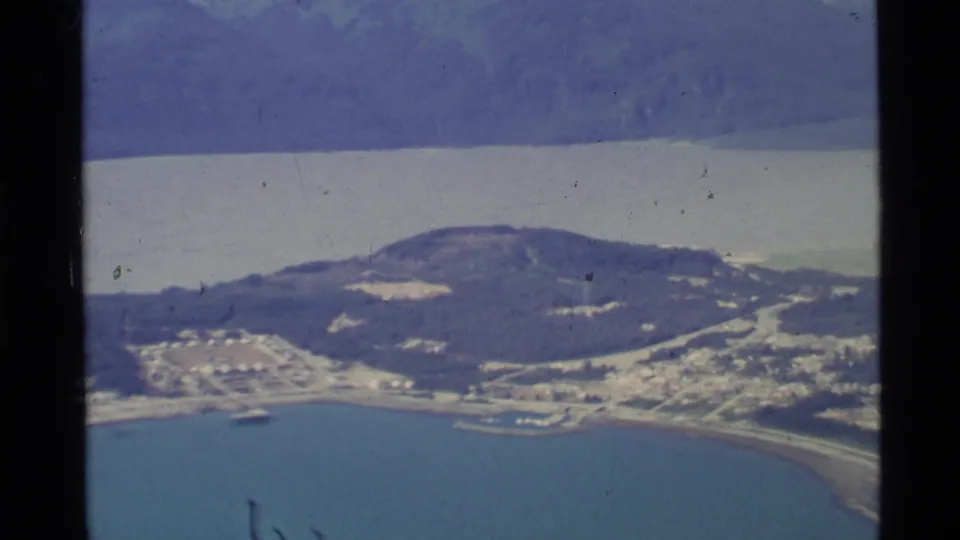 Aerial 8mm frame over coastal Alaska with a bay in the foreground, a wooded hill, small shoreline development, and hazy mountains beyond.