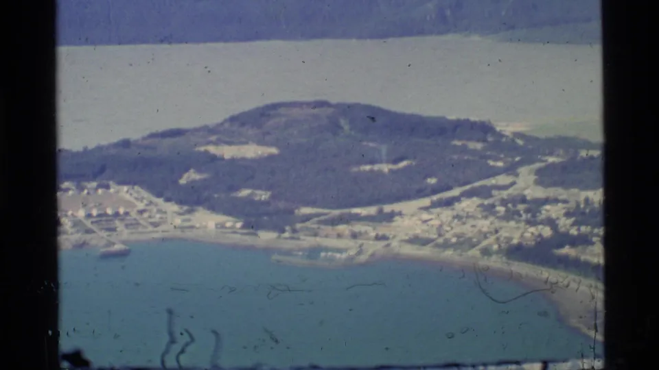Closer 1977 aerial view of an Alaskan shoreline settlement, with roads, waterfront structures, blue-green water, and a forested hill.