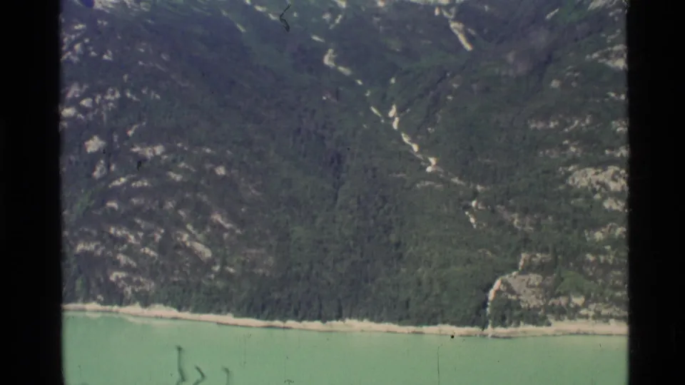 1977 8mm aerial frame showing coastal Alaska terrain, with pale green water, a narrow beach, dense forest, and steep mountain slopes.