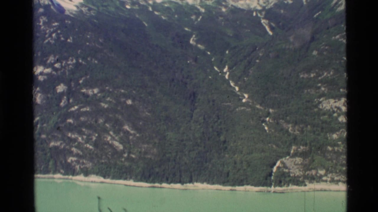 1977 8mm Alaska travel footage showing a turquoise lake bordered by forested mountains and lingering snow on higher slopes.