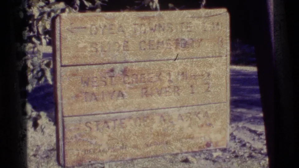 Soft 8mm frame of the Alaska directional sign for Dyea Townsite, Slide Cemetery, and Taiya River, seen in outdoor light.