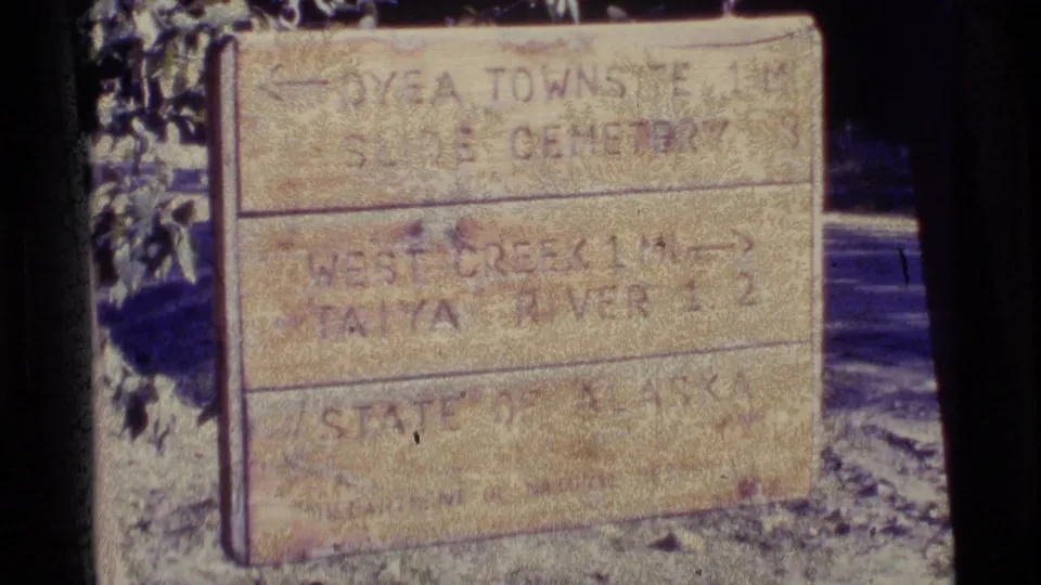 1977 Alaska 8mm frame showing a weathered sign with directions to Dyea Townsite, Slide Cemetery, West Creek, and Taiya River.