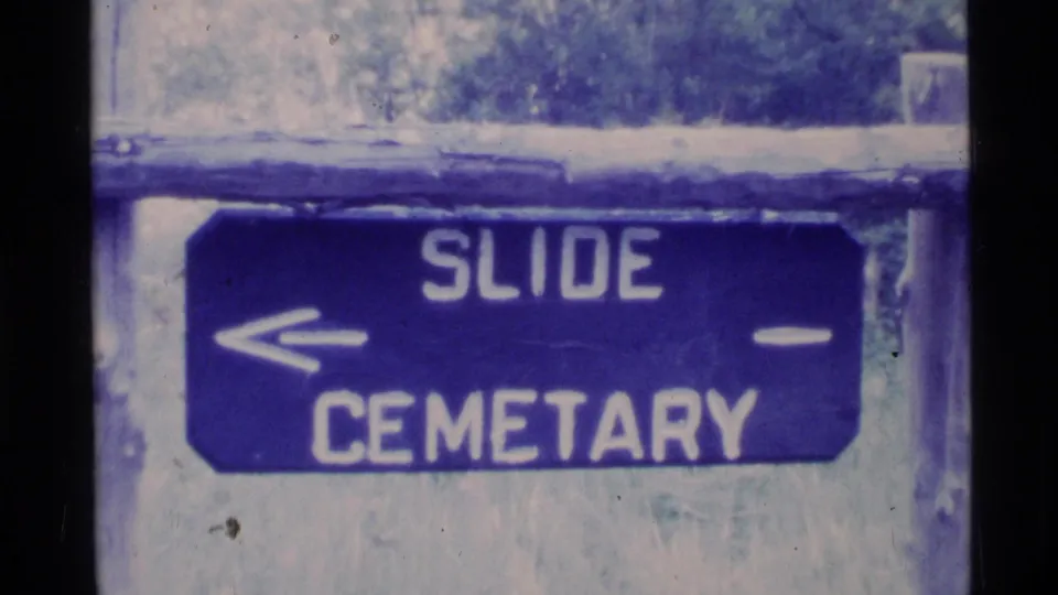 Blue directional sign in an Alaska home-movie frame, reading Slide Cemetary with a left arrow beside a wooded background.