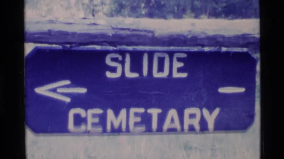 Close 8mm frame of a blue Slide Cemetary sign, with white lettering and a left arrow filling most of the view.