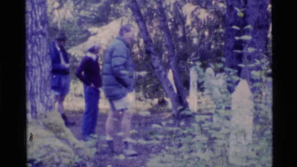 1977 Alaska 8mm frame showing visitors in casual clothing walking among trees and old cemetery markers.