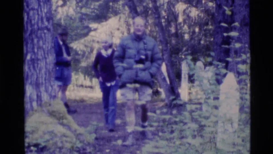 1977 Alaska home-movie frame with people walking through a forested cemetery as grave markers appear beside the path.