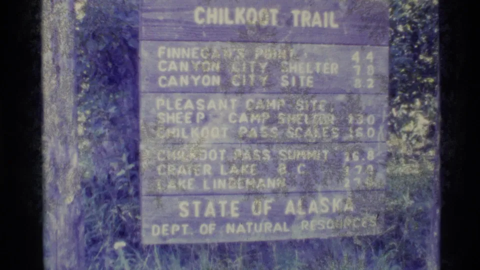1977 Alaska home-movie frame holding on a weathered Chilkoot Trail distance sign with visible 8mm grain and color fading.