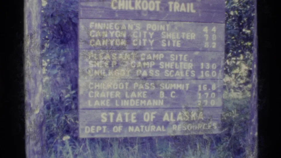 Final 1977 Alaska frame of the Chilkoot Trail wooden sign, showing public trail information and surrounding greenery.