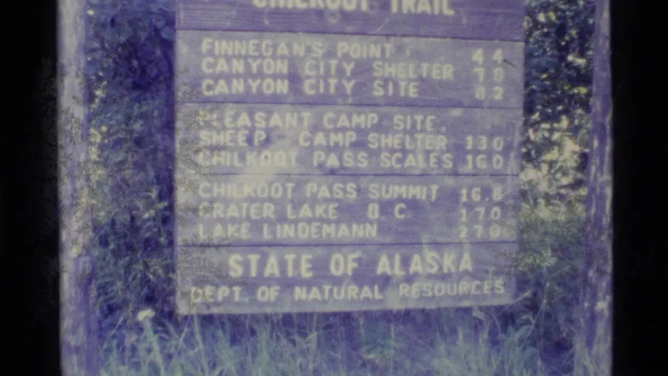 1977 8mm close-up of the lower Chilkoot Trail sign, showing camp, shelter, summit, and lake distance entries.
