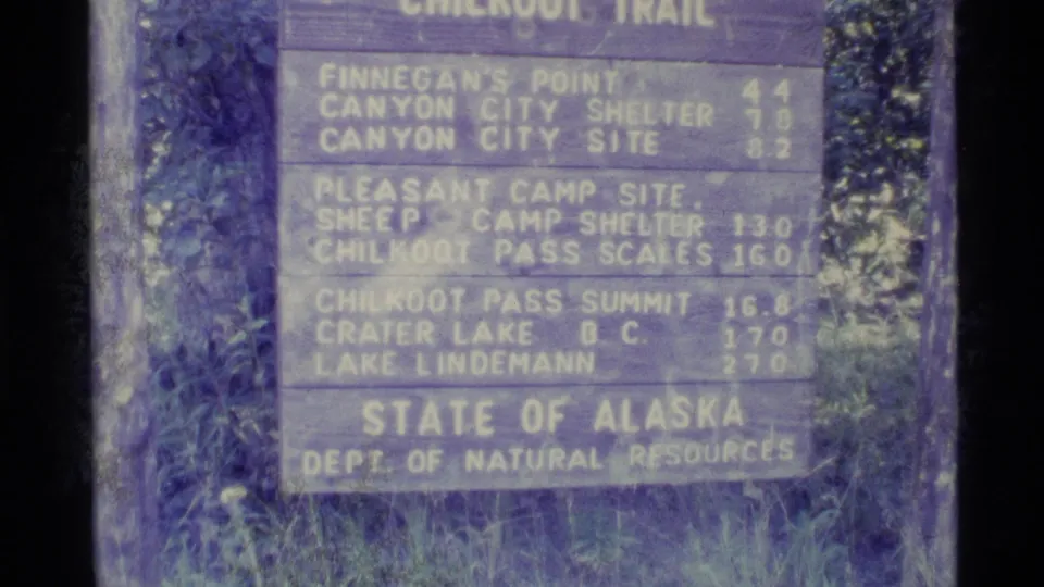 Soft, color-shifted 8mm frame showing the Chilkoot Trail distance sign in Alaska with official State of Alaska wording.