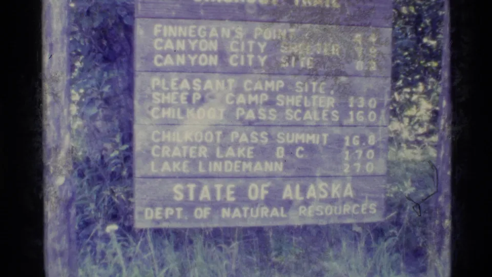 1977 Alaska home-movie frame of a weathered trail sign listing points including Chilkoot Pass Summit and Lake Lindemann.