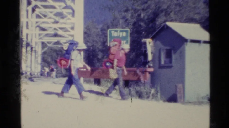 1977 Alaska 8mm frame showing two hikers with large backpacks walking near a small building and a visible Taiya sign.