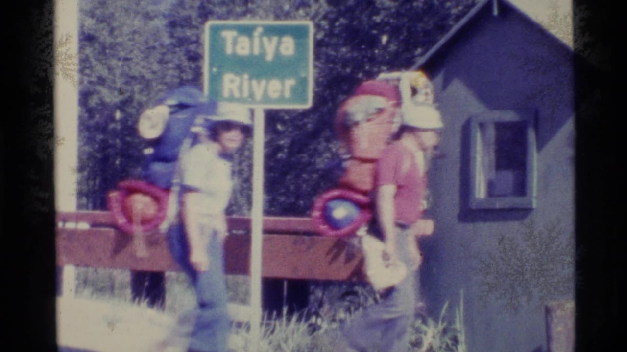 Two backpackers pass a green Taïya River sign beside a red railing and small structure.