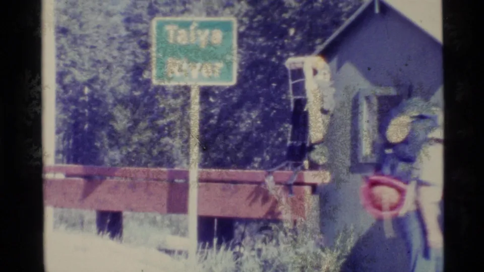 1977 Alaska frame of a hiker with a large backpack passing the Taïya River sign beside a trail structure.