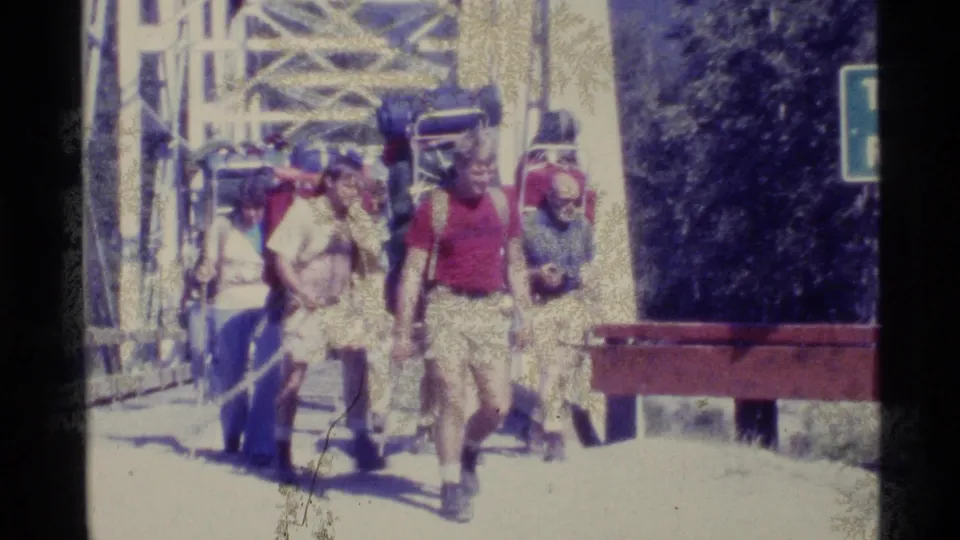 1977 Alaska home-movie frame showing a group of backpackers approaching or crossing near a metal truss bridge.