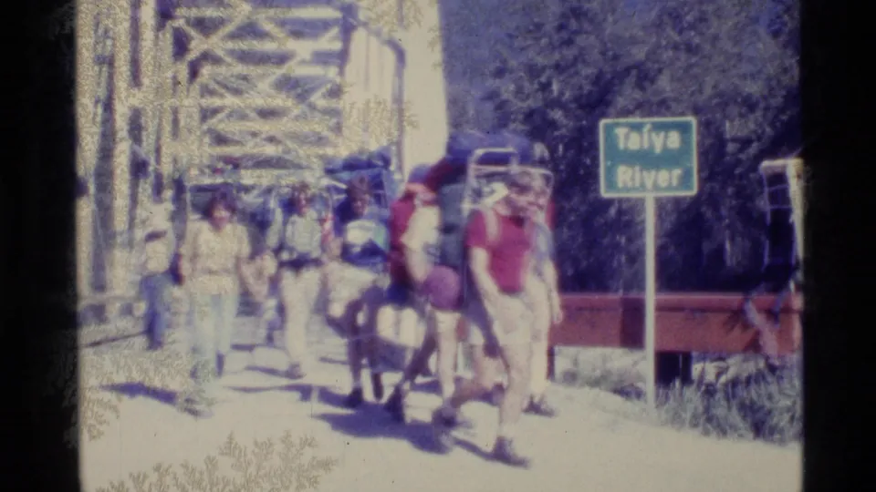1977 Alaska frame showing backpackers moving beside the Taïya River sign, red railing, and metal bridge structure.