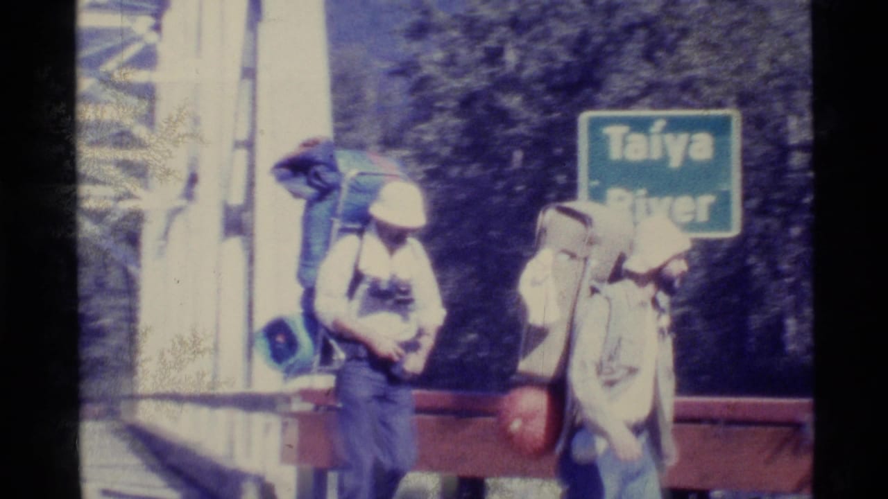 Two hikers with large packs pass a green Taïya River sign in 1977 Alaska home-movie footage.