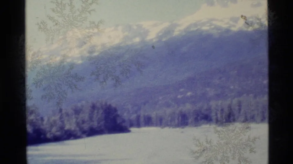 A wide 1977 Alaska home-movie view shows snow, trees, and mountain ridges in soft blue exposure.
