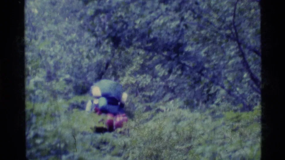 1977 Alaska home-movie frame of a hiker with a large pack moving through mossy forest under soft handheld focus.
