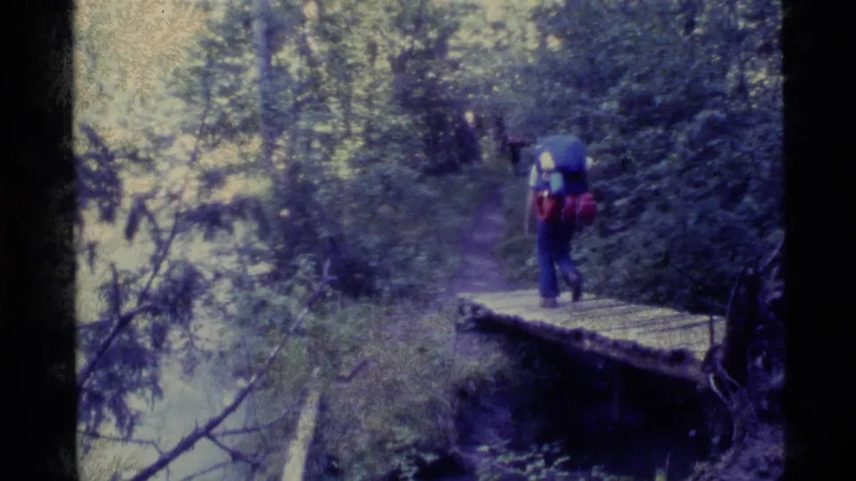 A backpacked hiker in blue and red clothing crosses a small wooden bridge on an Alaska forest trail, filmed on 8mm in 1977.