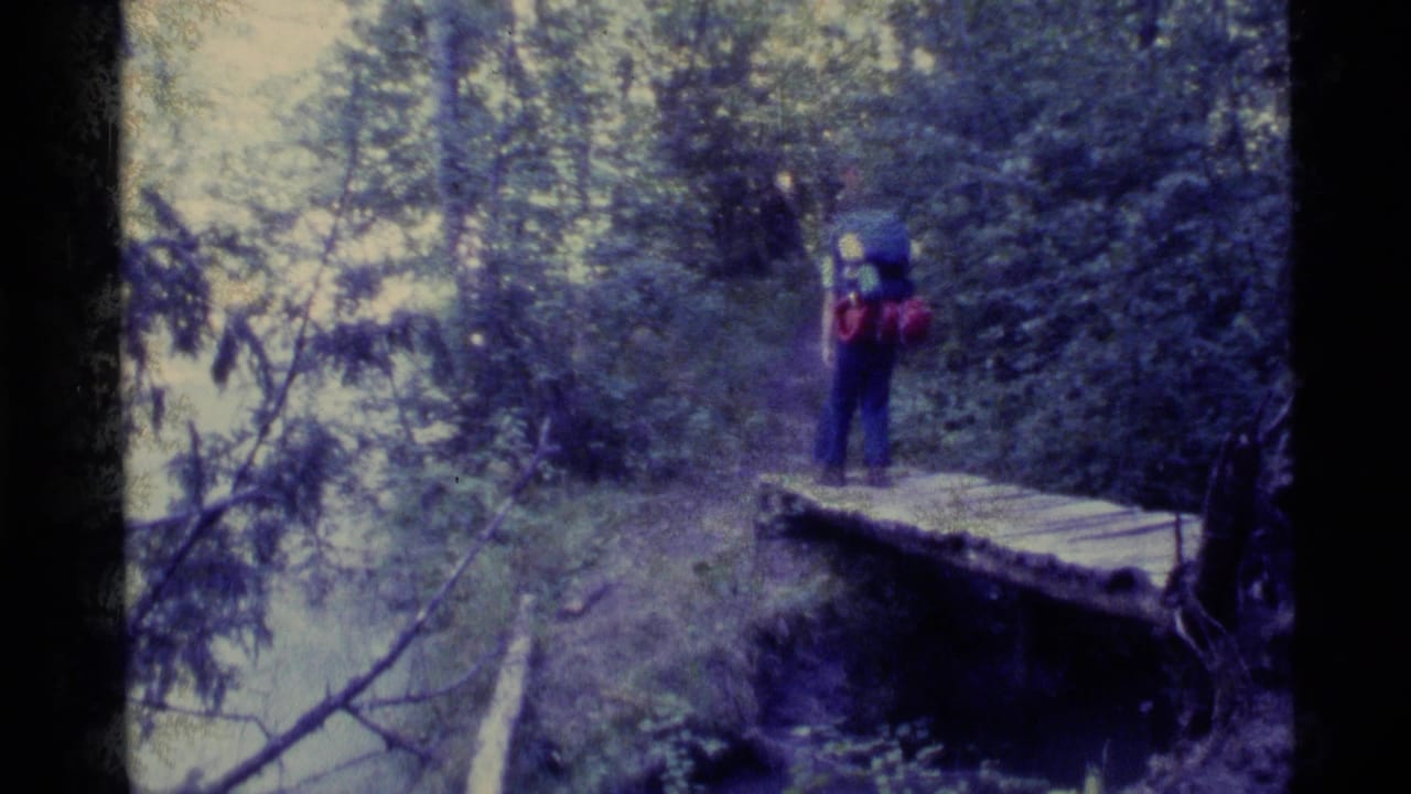 A backpacker crosses a small wooden bridge over water on a dense Alaska forest trail in 1977 home-movie footage.