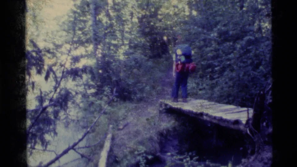 A backpacker crosses a small wooden bridge over water on a dense Alaska forest trail in 1977 home-movie footage.