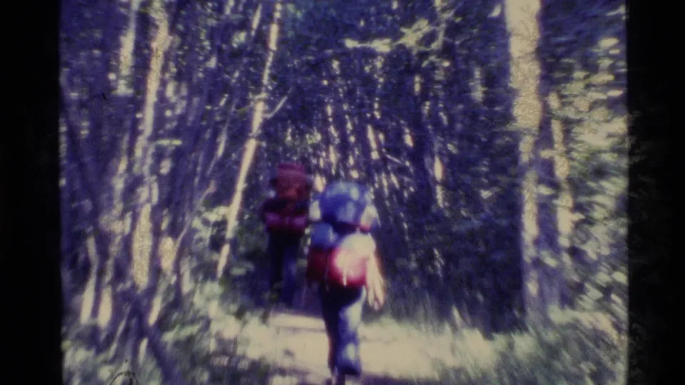 Two backpackers move away from the camera on a narrow forest path in 1977 Alaska home-movie footage.