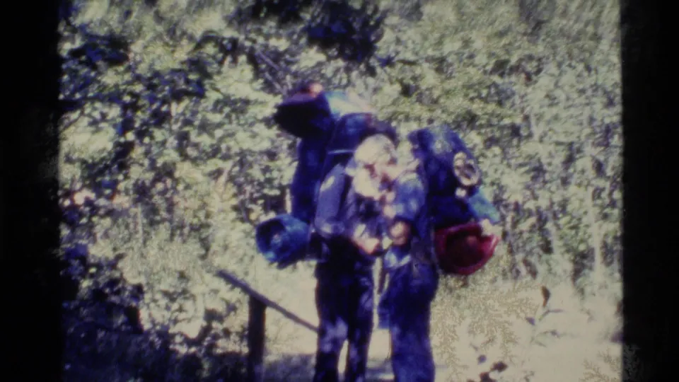 Closer 8mm frame of backpackers carrying external-frame packs near the wooded trail after the bridge crossing.