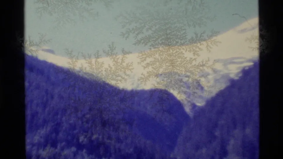 Wide Alaska landscape frame showing snowy peaks, dark evergreen hills, and pale sky in soft, grainy 1977 8mm footage.