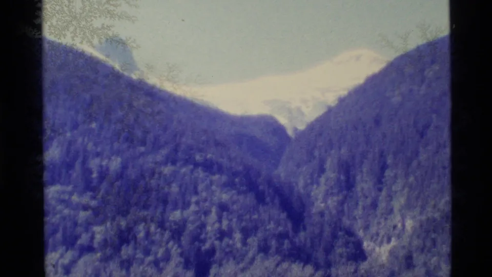 1977 Alaska home-movie frame with a distant snow ridge, evergreen-covered mountains, and a dark film-gate divider along the left edge.