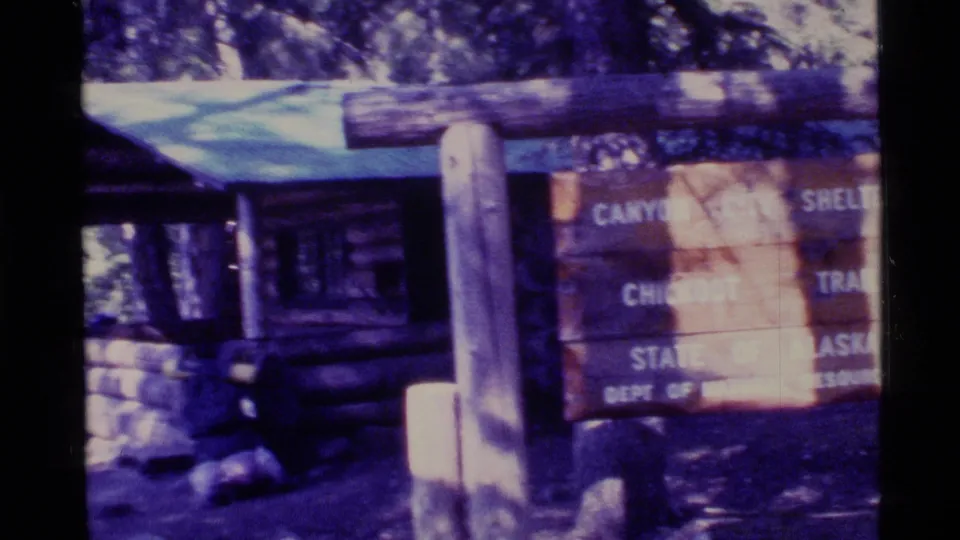 1977 Alaska frame showing the Canyon City Shelter area, with a log structure, wooden post, forest shade, and partial trail sign.