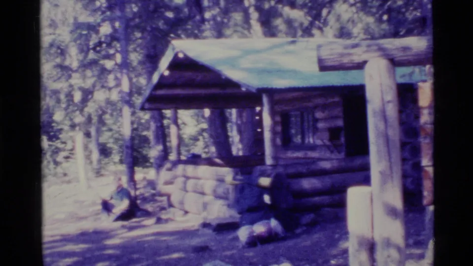 1977 Alaska 8mm frame showing a rustic log shelter in forest shade, with a roof overhang, stonework, and trail signpost nearby.