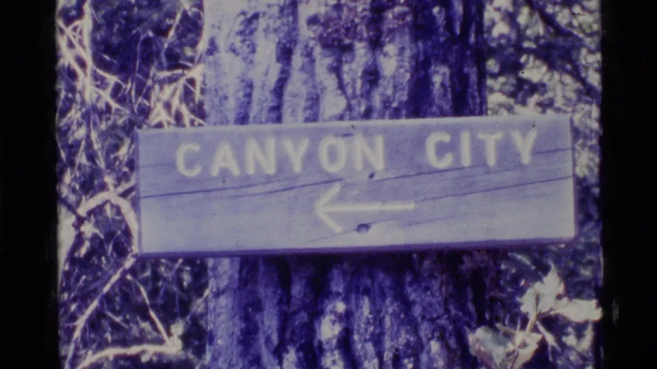 1977 Alaska 8mm frame showing a weathered wooden Canyon City sign with an arrow attached to a tree among forest growth.