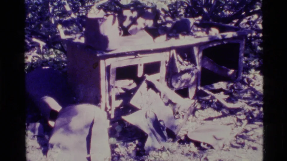 1977 Alaska home-movie frame showing a damaged wooden structure or cabin remains surrounded by forest and scattered debris.