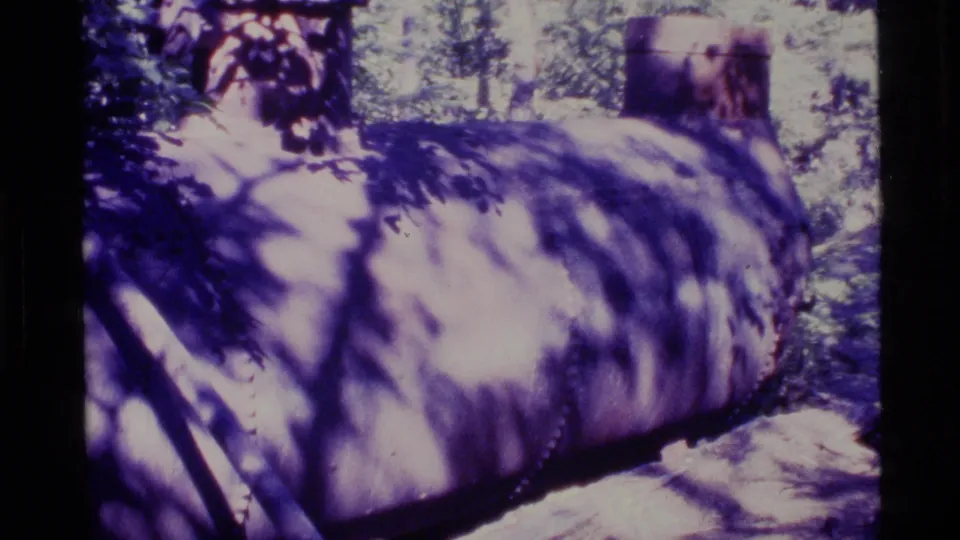 1977 Alaska 8mm frame showing a rusted cylindrical tank or pipe section on the ground beneath trees and dappled shadows.